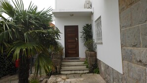 Un edificio blanco con una puerta marrón, escalones de piedra y un pequeño jardín con palmeras.