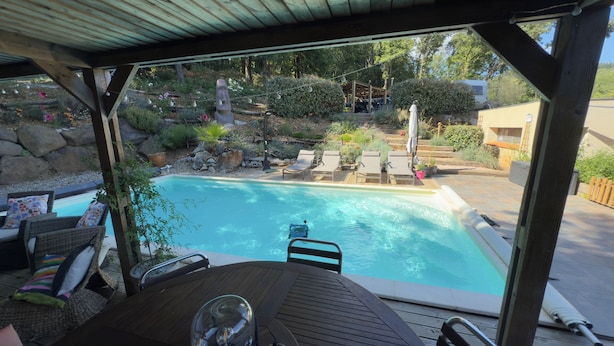 Ein Poolbereich mit Überdachung, Liegestühlen und einem Tisch, umgeben von einem angelegten Garten mit diversen Pflanzen und einem kleinen Wasserfall.