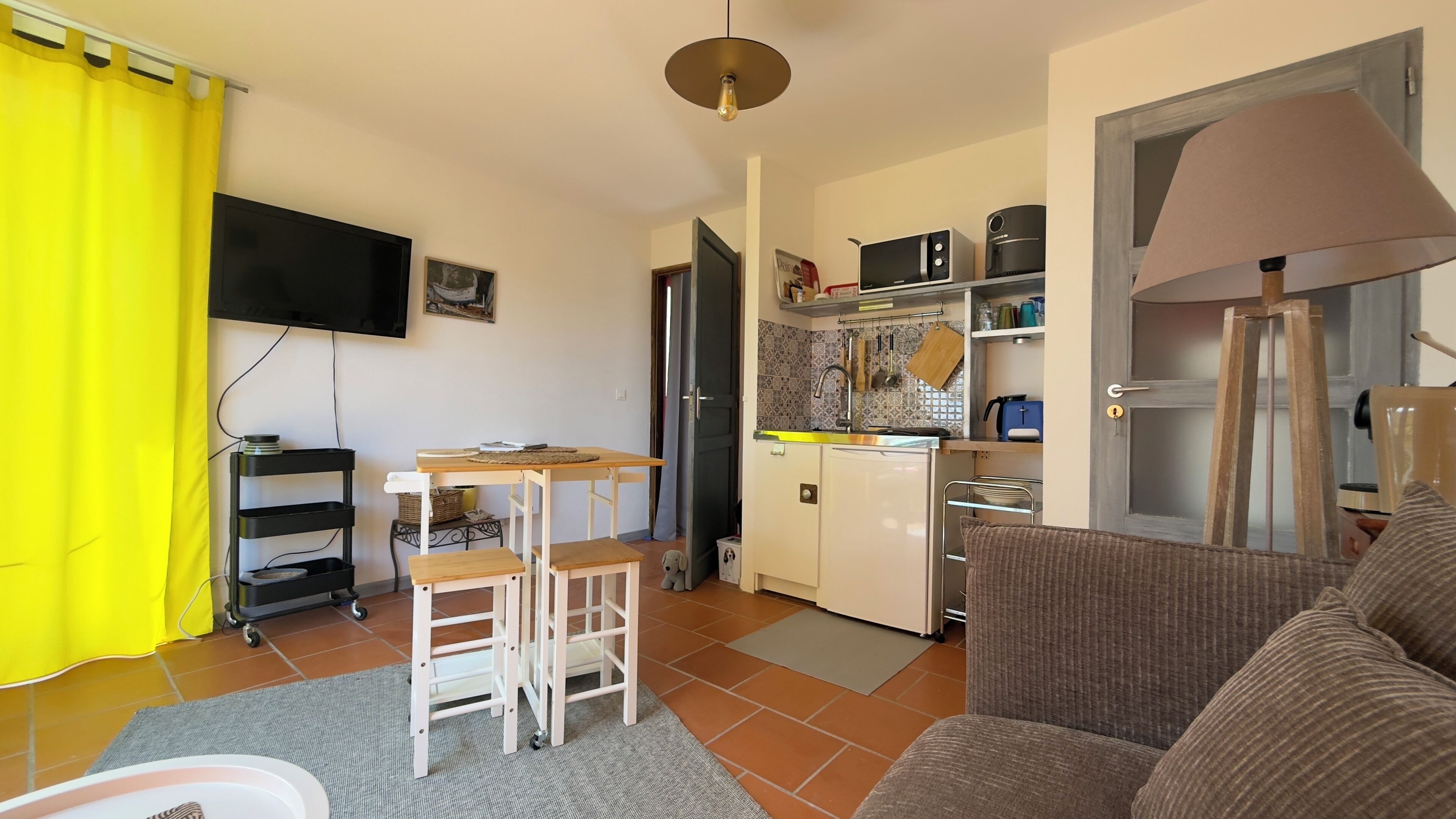 A compact living space with a kitchenette, a dining area with a small table and chairs, a sofa, and a TV mounted on the wall.