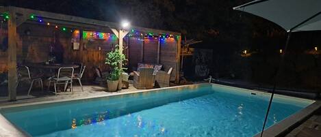 A swimming pool with a wooden deck, outdoor seating, and string lights.