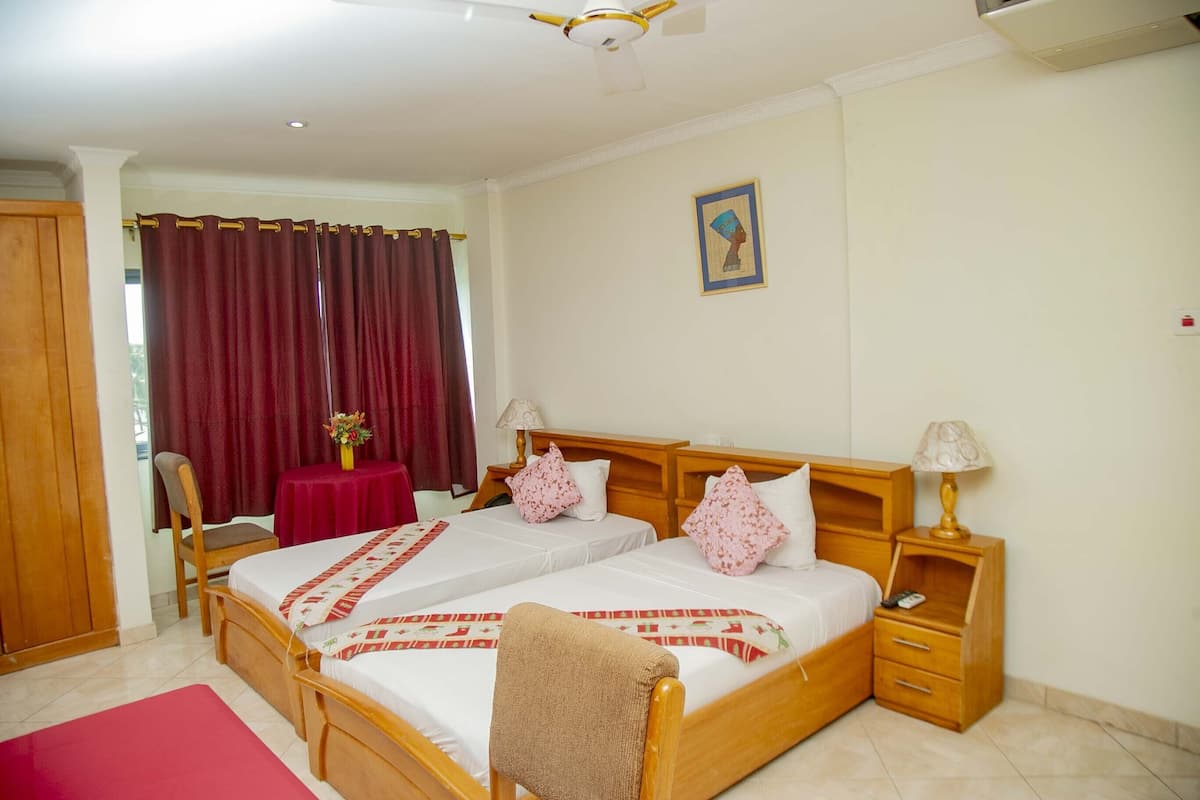 A hotel room with two beds, a wooden nightstand, a chair, a table with a vase of flowers, and a painting on the wall.