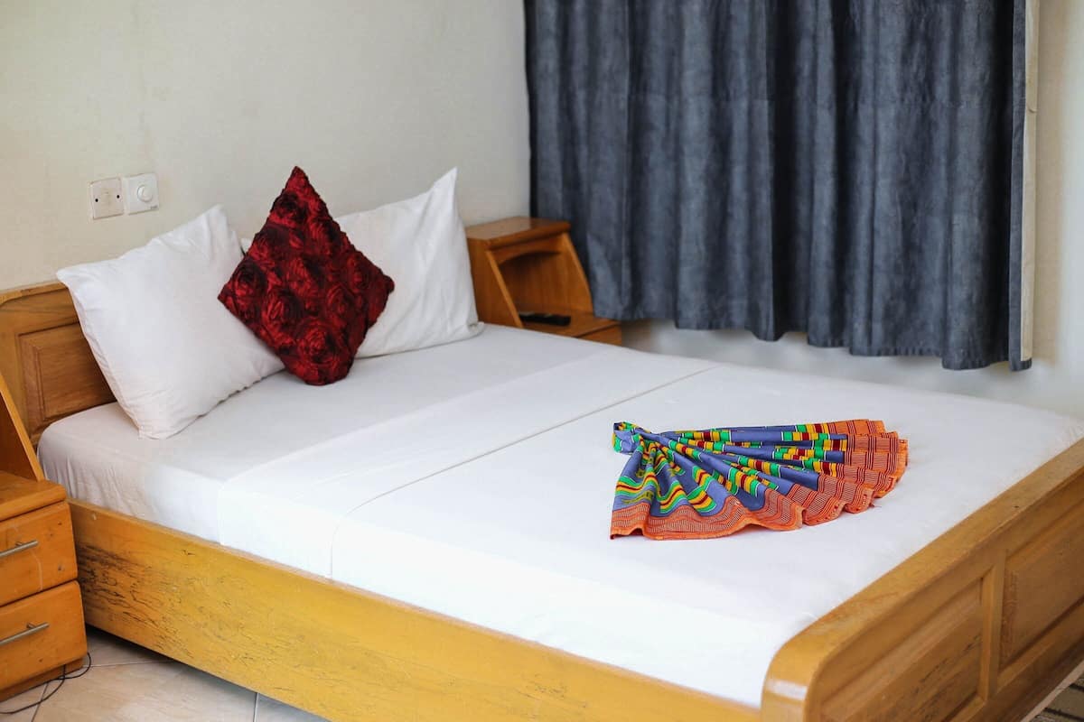 A neatly made bed with white sheets, a red pillow, and a colorful blanket. There is a wooden nightstand beside the bed with a remote control on it.