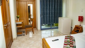 Una camera d'albergo con un letto, una scrivania con una sedia, un piccolo frigorifero, un comodino e una finestra con tende.