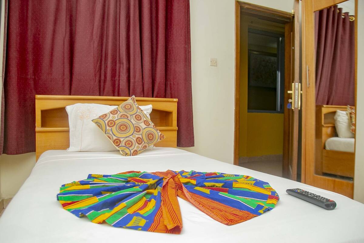 A hotel room with a bed, a colorful blanket, a remote control, and a door leading to a bathroom.