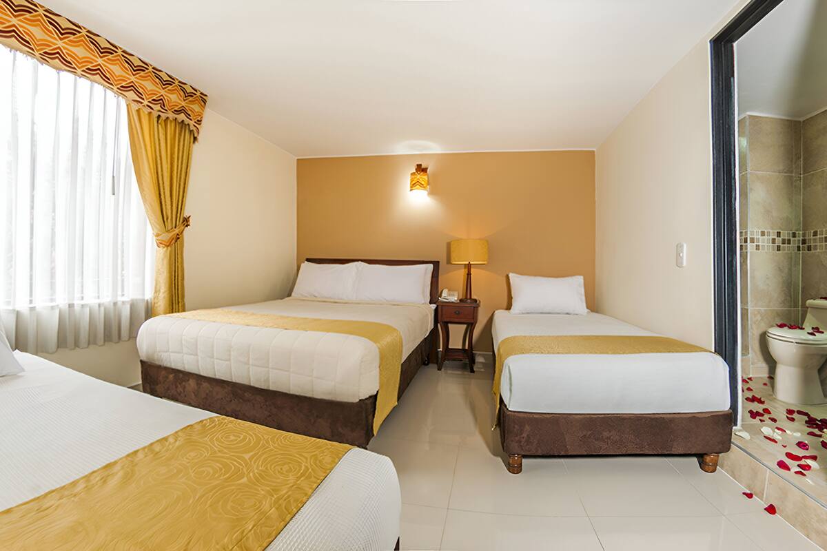 A hotel room with two beds, a bedside table with a lamp, a bathroom with a shower, and a toilet with rose petals.