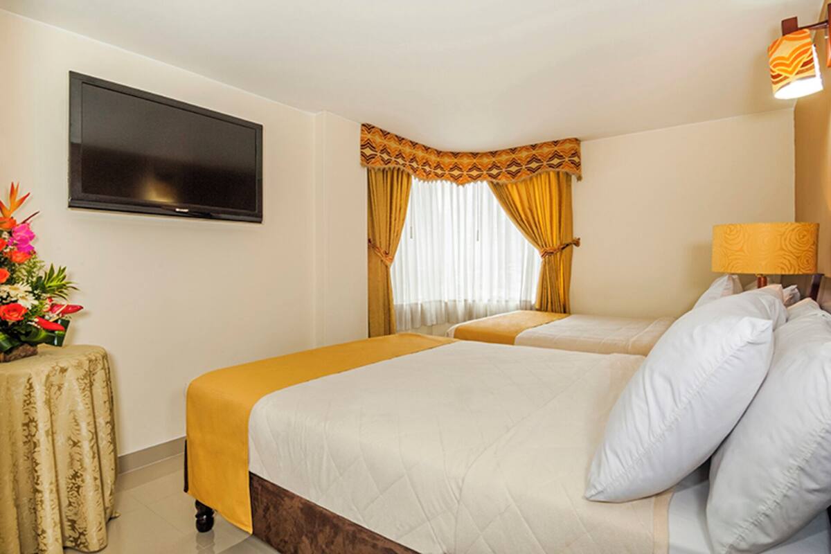 A hotel room with two beds, a flat-screen TV on the wall, a small table with a floral arrangement, and a window with curtains.
