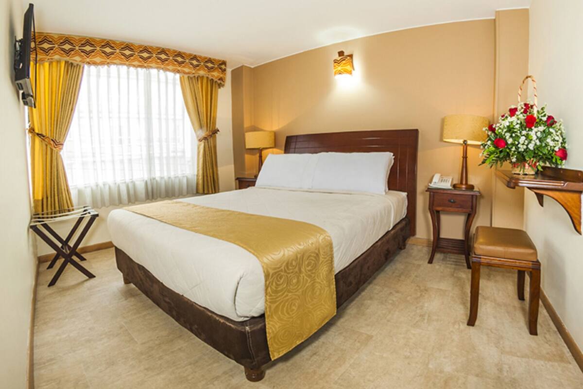 A hotel room with a large bed, a bedside table with a lamp, a small table with a telephone, a chair, and a wall-mounted shelf with a flower arrangement.