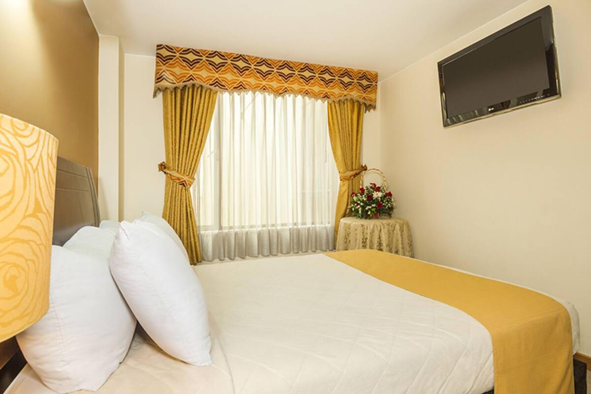 A hotel room with a bed, a TV mounted on the wall, and a window with curtains.