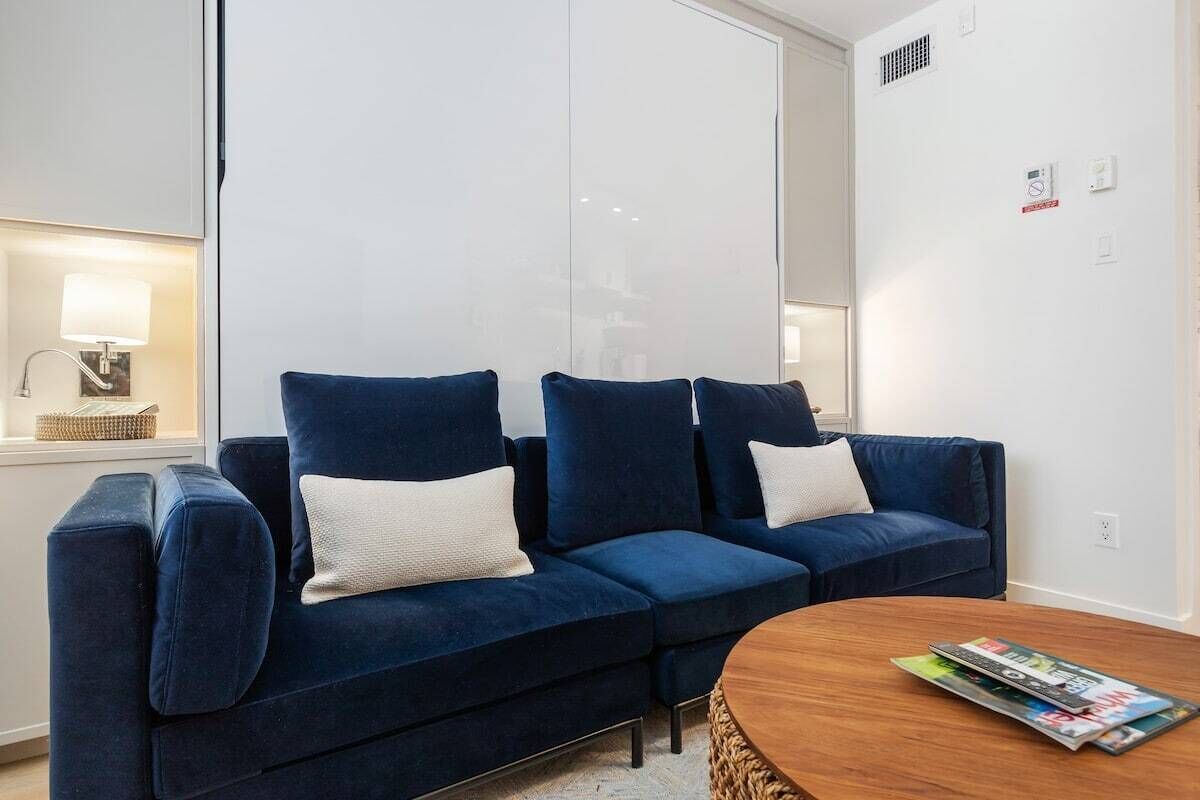 A modern living room with a dark blue sofa set, a round wooden coffee table, and a large window with blinds.