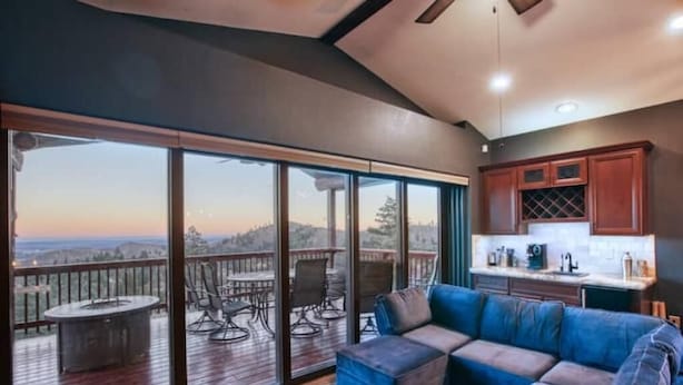 Un espacio habitable amplio con un gran ventanal corredizo que da a una terraza exterior, un sofá modular confortable y una cocina con armarios de madera.