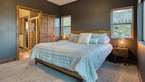 Un dormitorio con una cama grande, cabecera de madera, dos mesitas de noche con lámparas y vistas al exterior a través de una ventana.