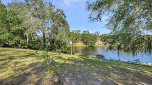 A serene lakeside setting with a grassy area, trees, and a dock extending into the water. - Stunning Lake House w/ Heated Pool + Ping Pong! (Lutz)