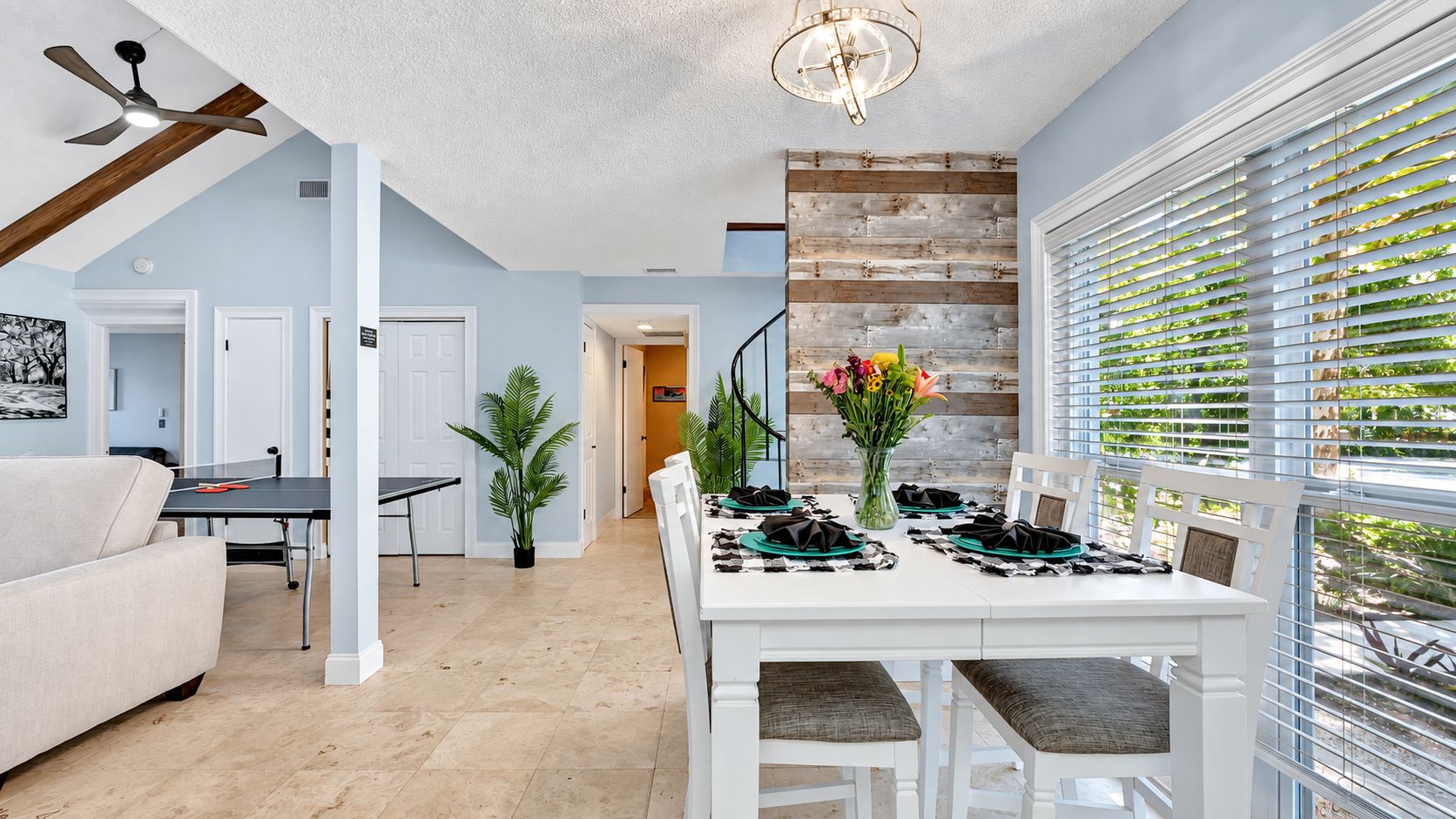 A dining area with a white table set for four, a ceiling fan, and a view through the window blinds.