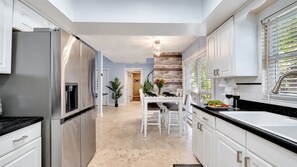 Une cuisine moderne dotée d’appareils électroménagers en acier inoxydable, un coin repas avec des meubles blancs et une vue sur l’extérieur grâce à une fenêtre équipée de stores.
