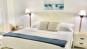 Un lit bien fait, recouvert d’une couverture bleue, avec deux lampes de chevet, un tableau encadré au-dessus du lit et une table de chevet blanche dotée de tiroirs.