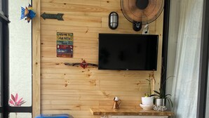 Une pièce de taille modeste avec un mur en bois, un téléviseur fixé au-dessus d’un meuble blanc, un barbecue bleu et une petite poubelle.
