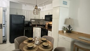 Une cuisine compacte avec des meubles blancs, un réfrigérateur noir et un four à micro-ondes. On y trouve une table à manger avec quatre chaises et une lampe sur le côté droit.
