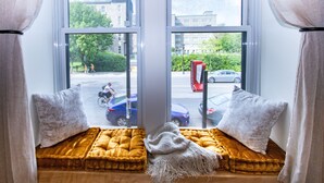 Asiento junto a la ventana con cojines y una manta, con vistas a una calle con coches y un ciclista.