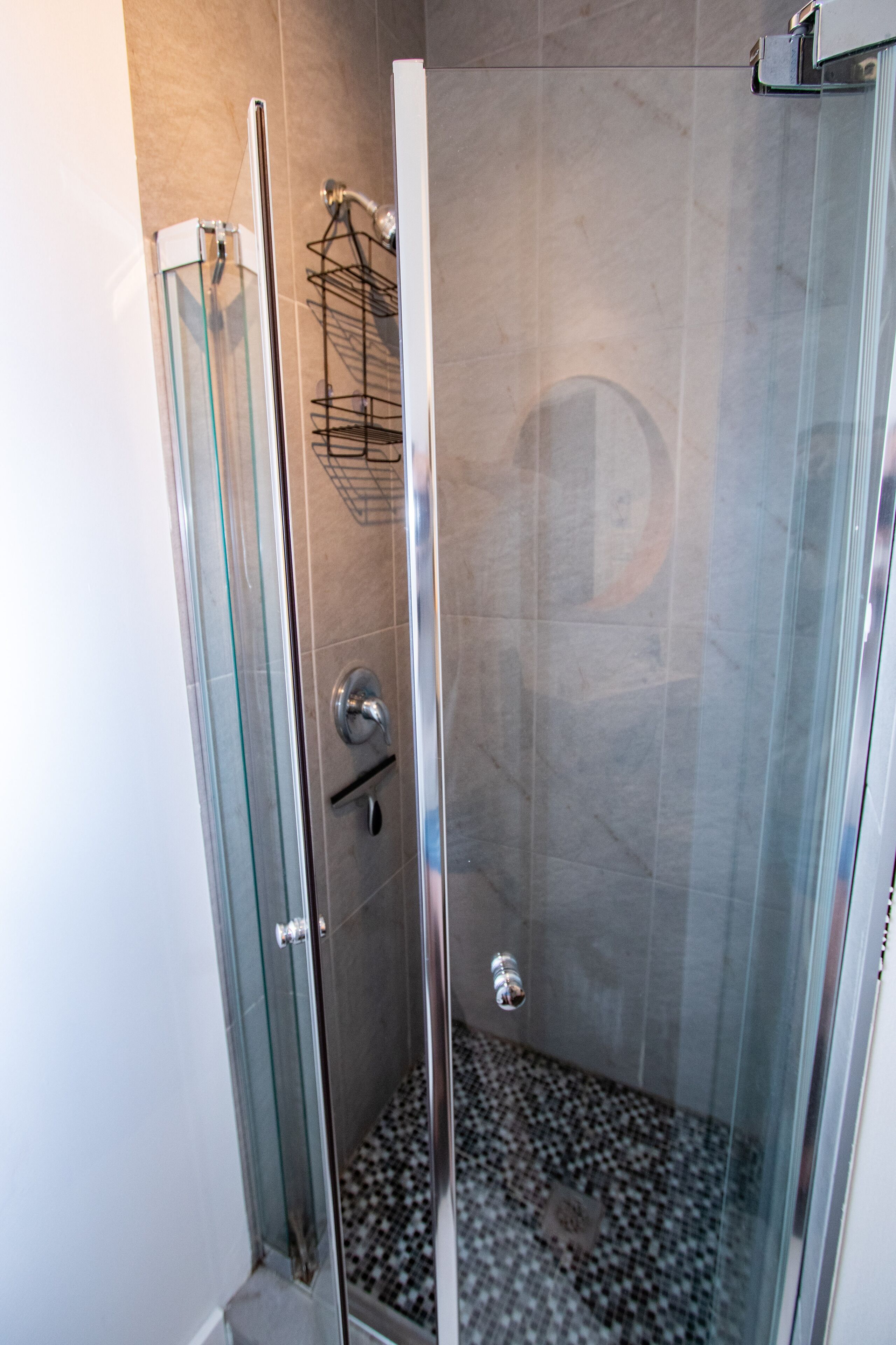 Une douche fermée par des parois de verre, avec un sol carrelé et une étagère en métal.