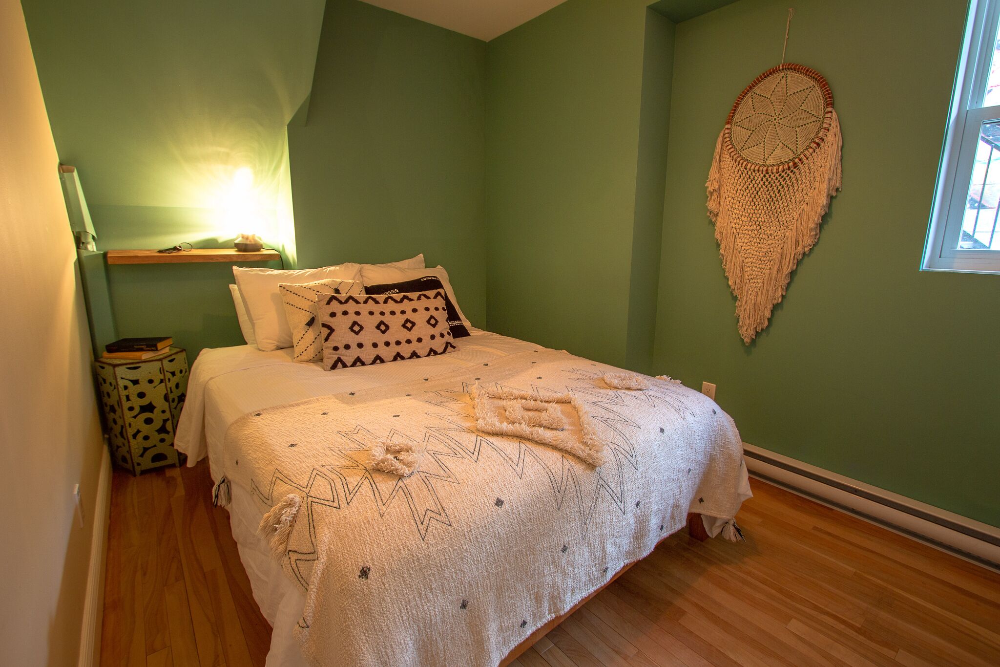Un lit bien fait, agrémenté d’oreillers à motifs et d’une décoration murale, dans une pièce aux murs verts et au plancher en bois.