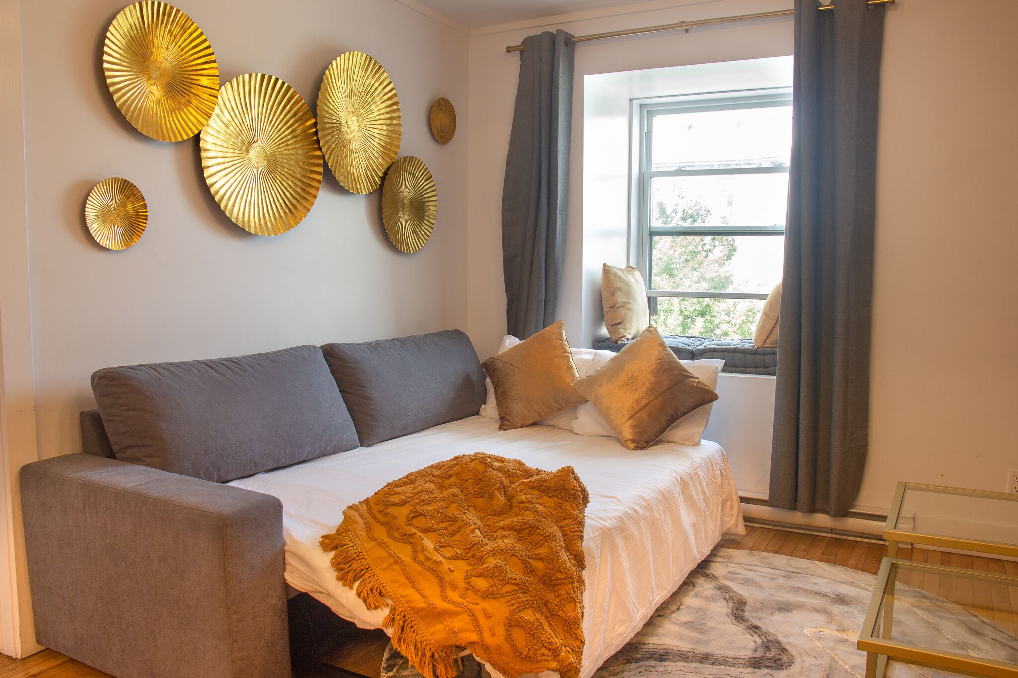 Un dormitorio moderno con un sofá gris, una cama blanca y detalles decorativos dorados en la pared.