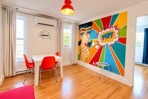 Ein Zimmer mit einem weißen Tisch, roten Stühlen, einem farbenfrohen Wandbild und einer roten Pendelleuchte.