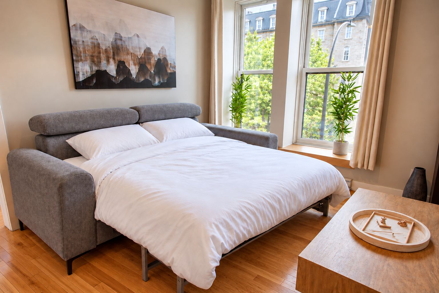 Ein modernes Schlafzimmer mit einem grau gepolsterten Bett, weißer Bettwäsche, einem Holznachttisch und einem Stadtbild an der Wand.