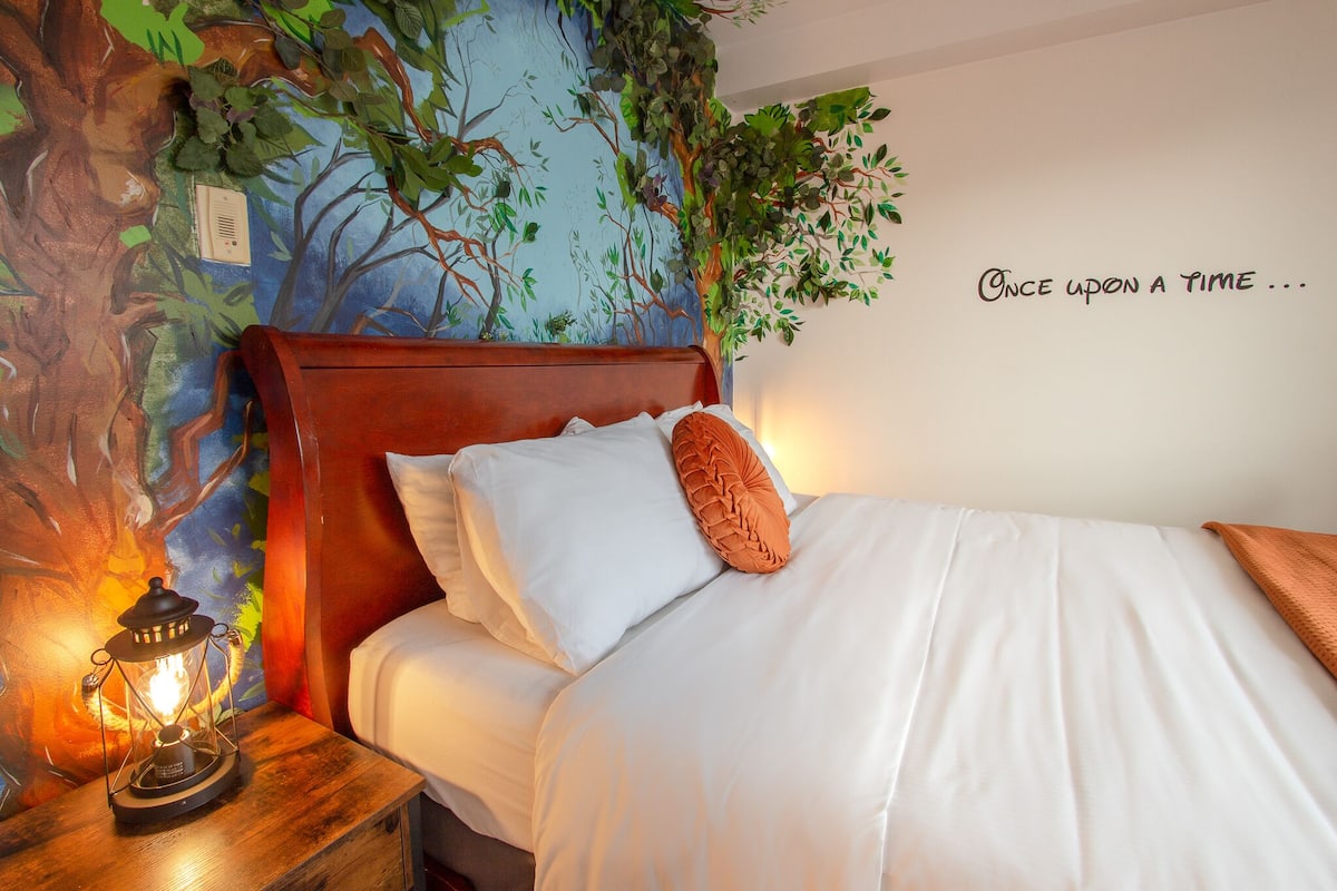 A neatly made bed with white linens and a single orange pillow, a wooden nightstand with a lantern, and a wall mural with greenery.