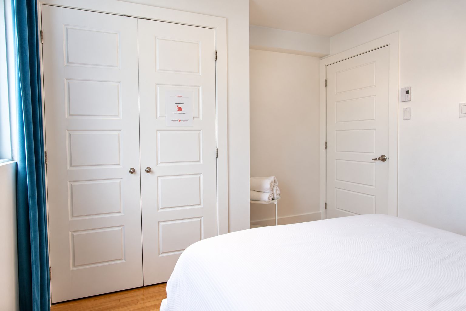 A hotel room with a bed, two white doors, a towel rack, and a thermostat on the wall.