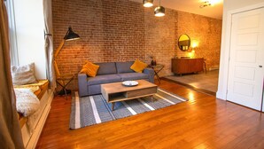 Un salón con sofá, mesa de centro y pared de ladrillo. Hay una puerta y una lámpara en la habitación.