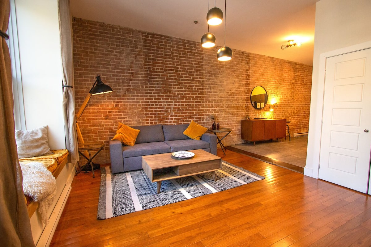A living room with a sofa, coffee table, and a brick wall. There is a door and a lamp in the room.