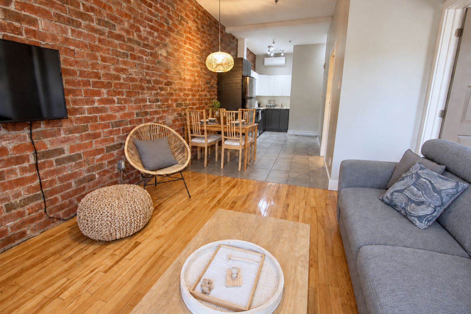 A living room with a brick wall, a flat-screen TV, a grey sofa, a round coffee table, and a dining area with wooden chairs and a hanging light fixture.