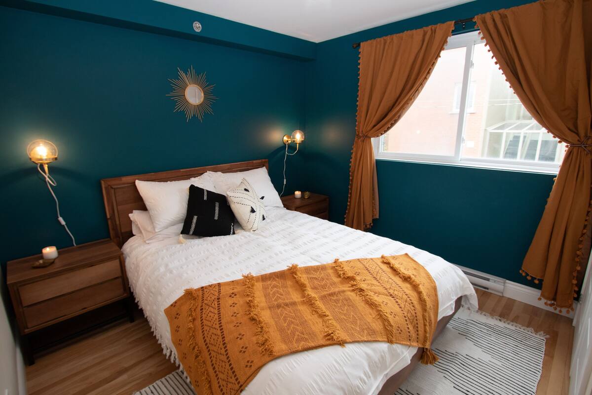 A bedroom with teal walls, a wooden bed, white bedding, a mustard-colored throw blanket, and a window with brown curtains.
