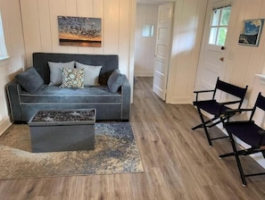 A living room with a grey sofa, patterned cushions, a coffee table, and a rug. There are two black chairs and a door in the background.