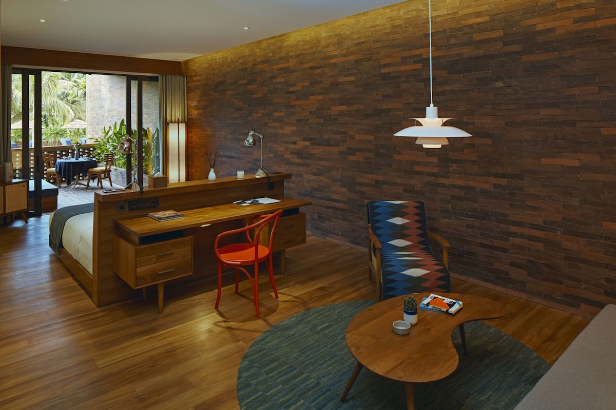 A modern hotel room with a wooden desk, a red chair, a sofa, a round coffee table, and a large window offering a view of the outdoors.