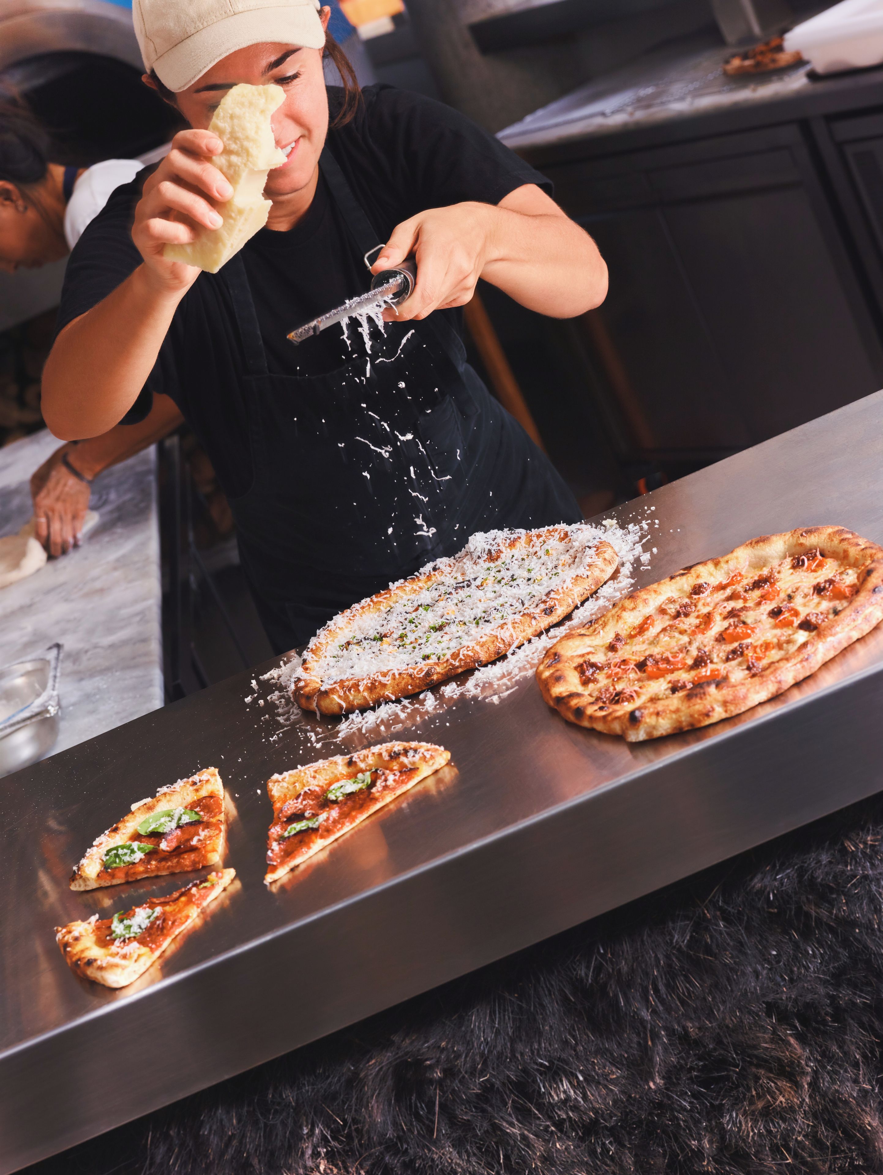 Iemand strooit geraspte kaas over een pizza op een metalen aanrecht.