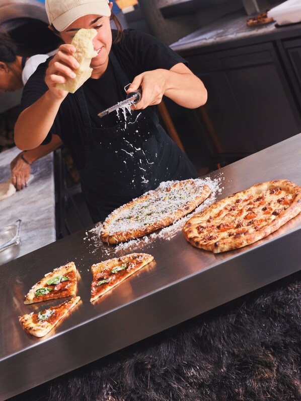 A person grating cheese over a pizza on a metal countertop.