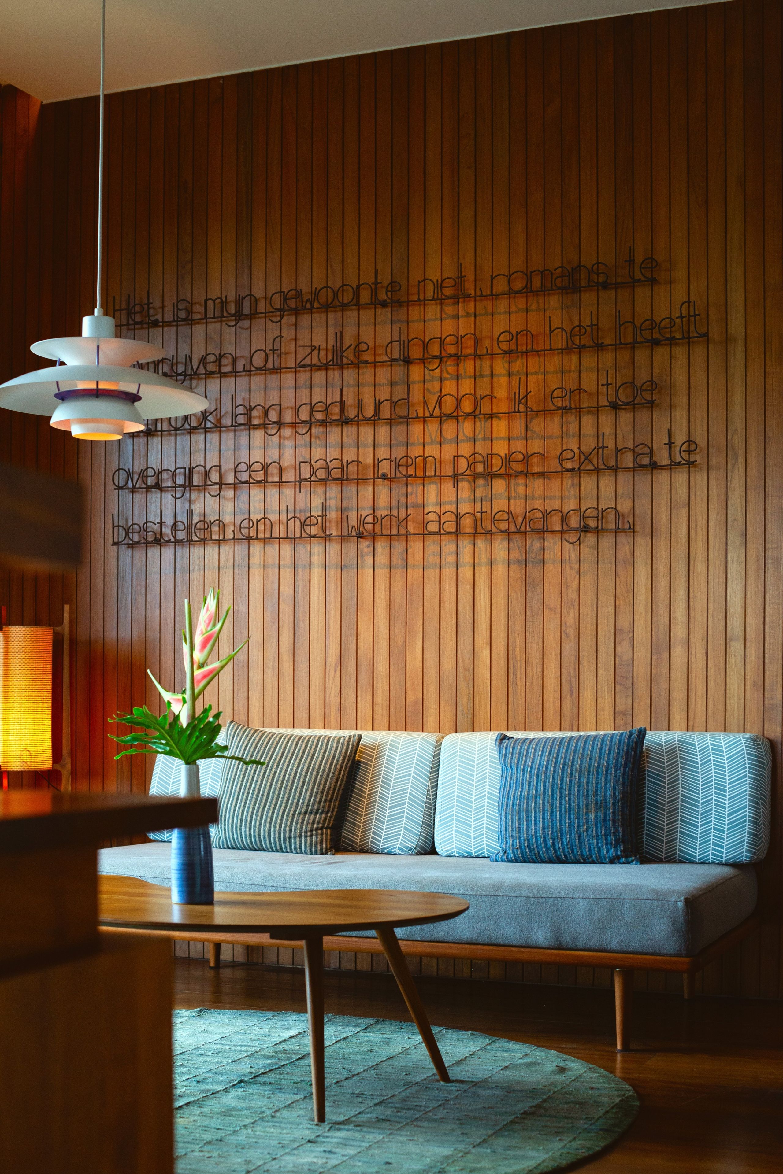 Un interior moderno con una pared de madera, un sofá con cojines estampados, una mesa redonda de madera y una lámpara colgante.