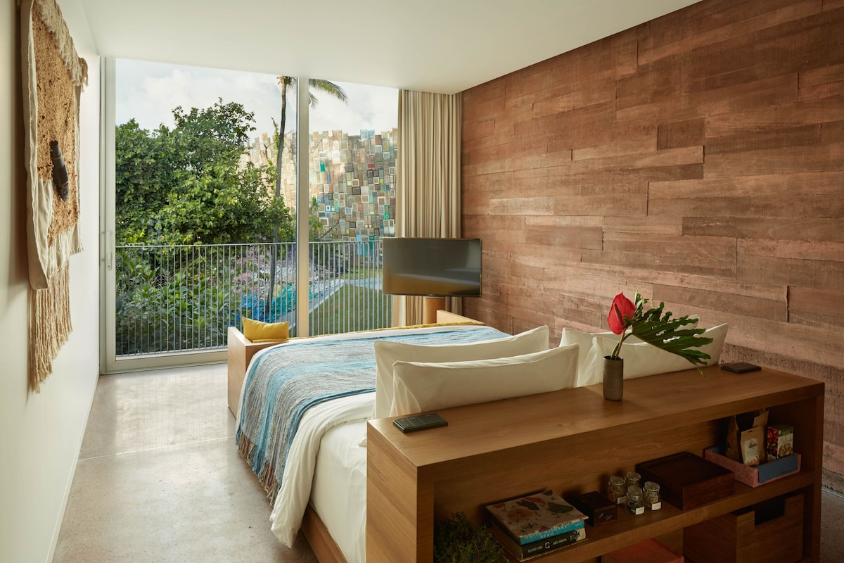 A bedroom with a bed, a TV, a wooden nightstand, and a balcony with a view of buildings and greenery.