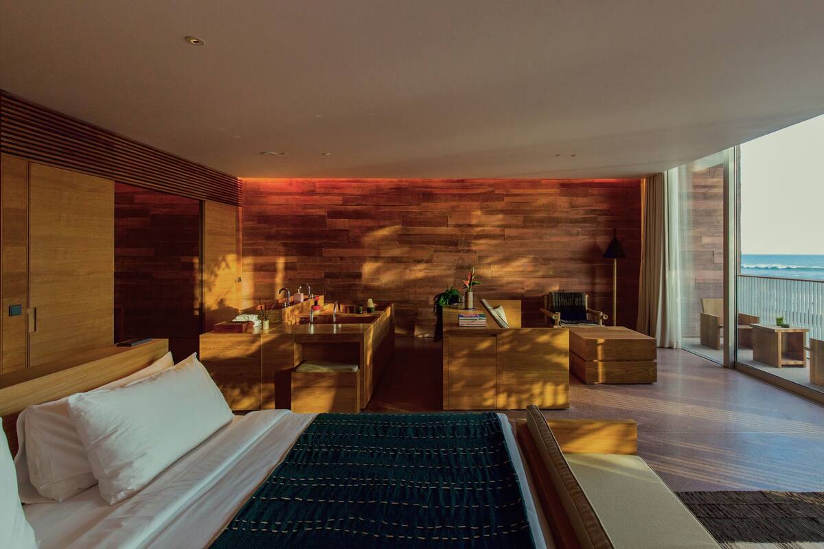 A modern bedroom with a large bed, a wooden wall, a balcony with a view of the ocean, and a seating area with a table and chairs.