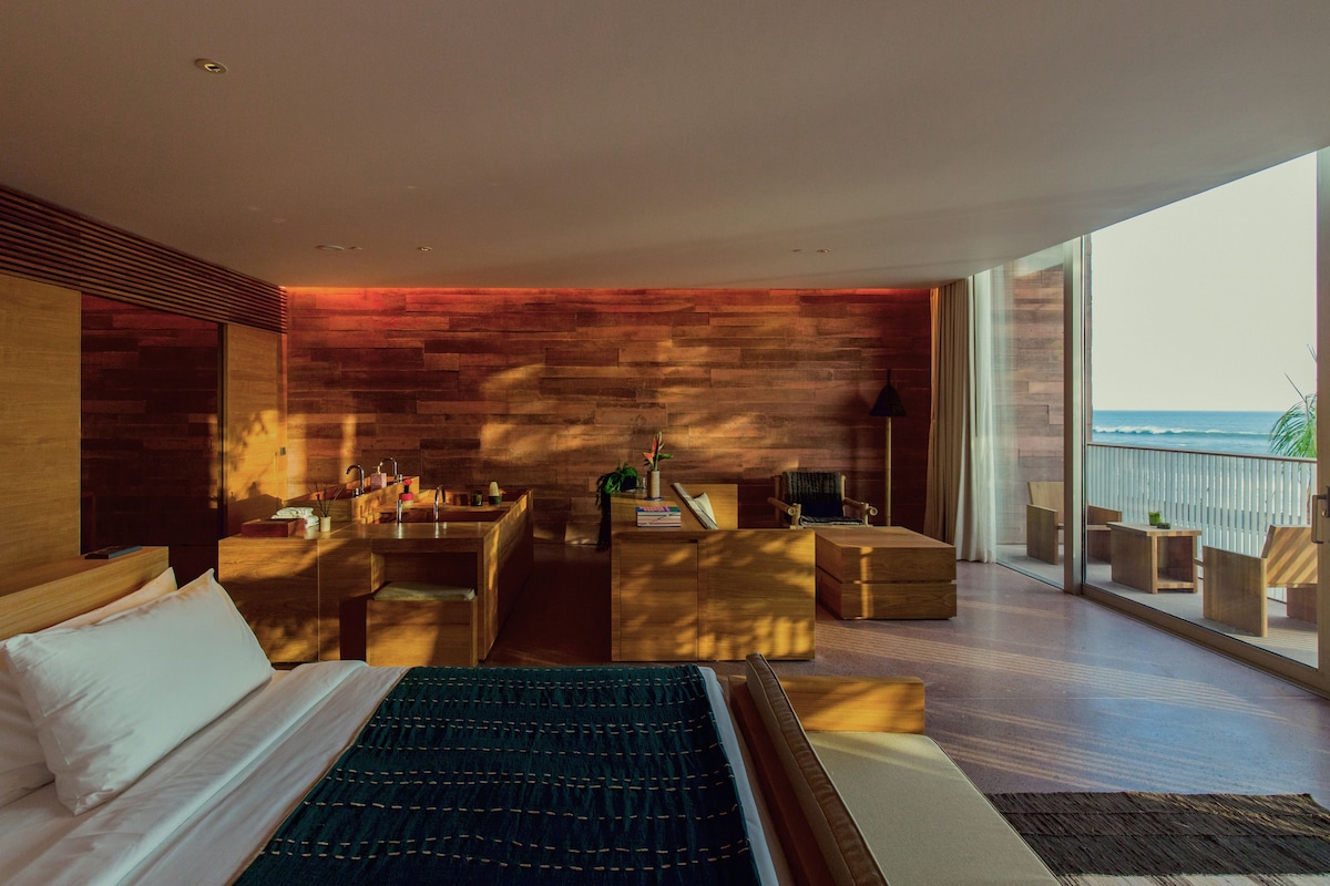 A modern bedroom with a large bed, a wooden wall, a balcony with a view of the ocean, and a seating area with a table and chairs.