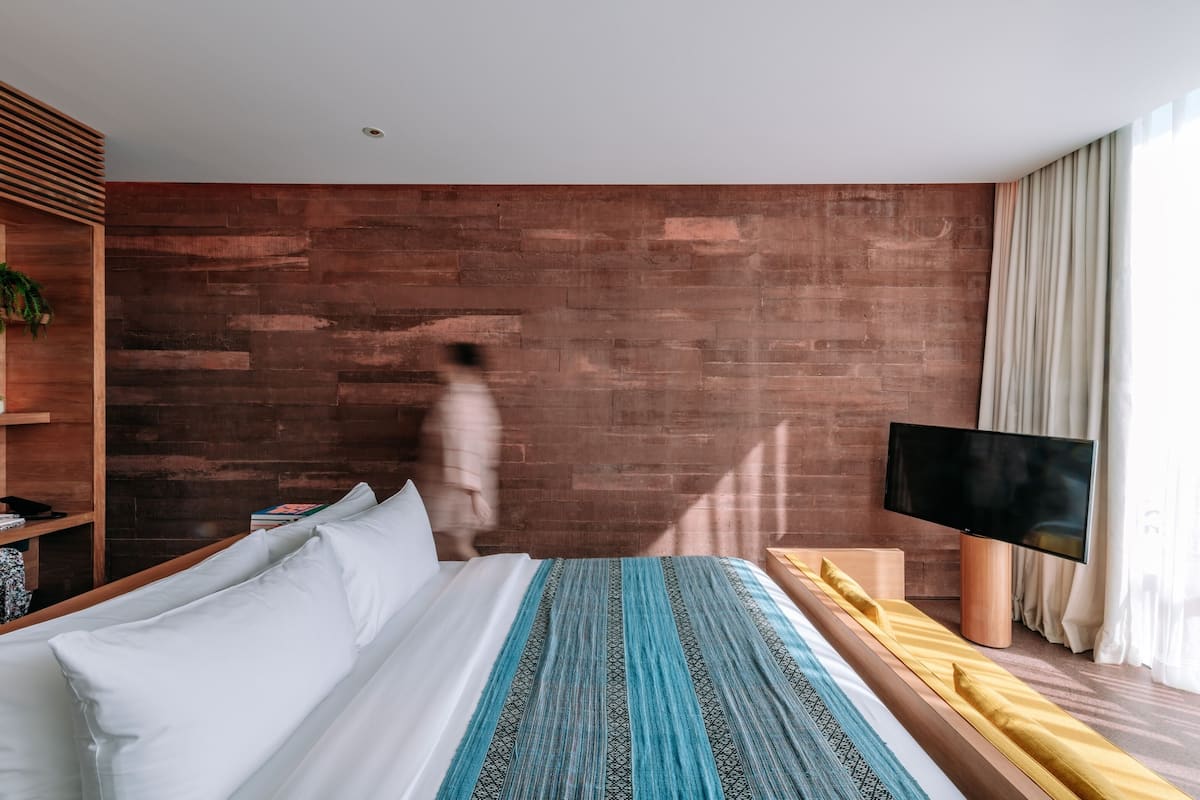 A modern hotel room with a wooden accent wall, a bed with white and patterned bedding, a TV, and a bedside table.