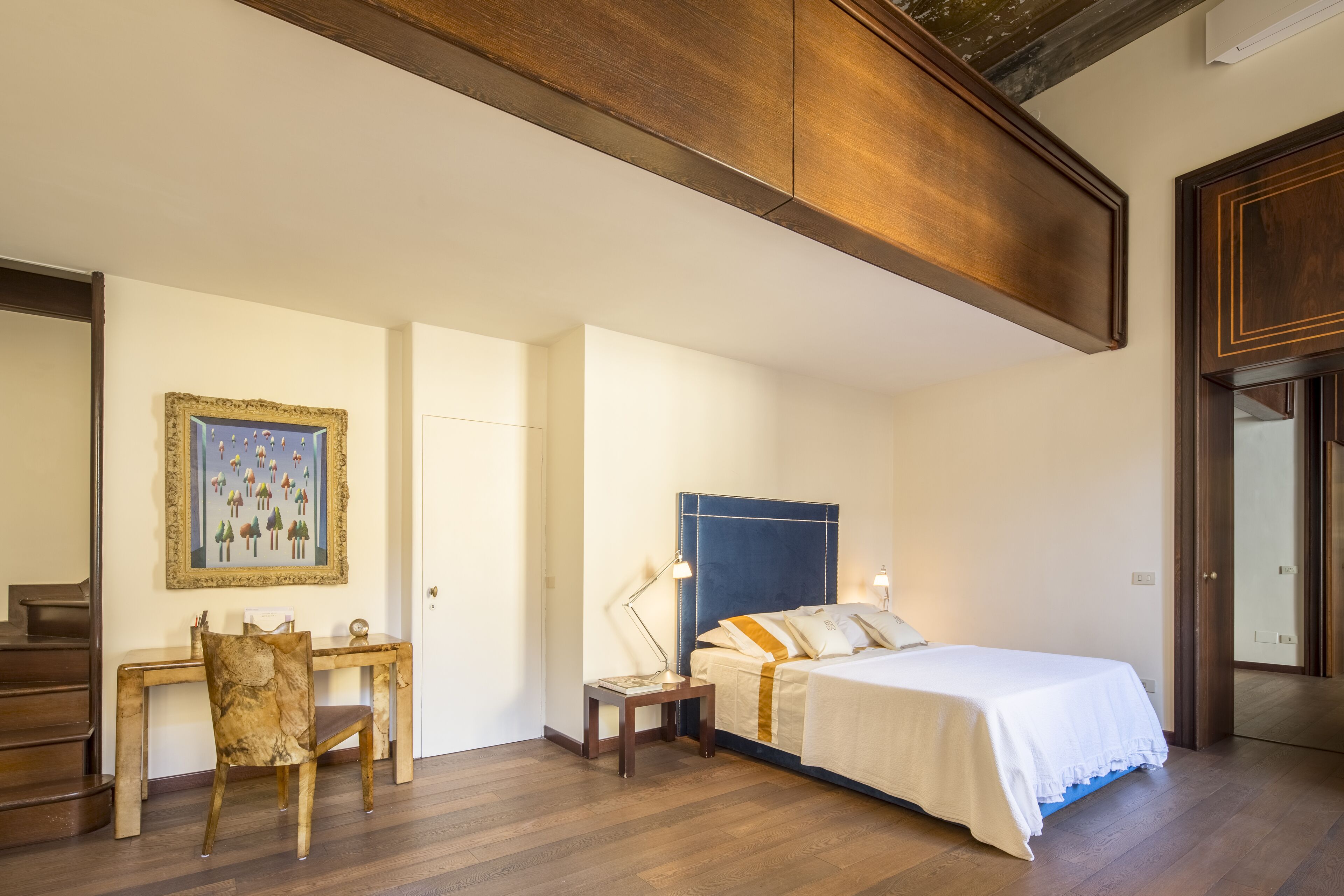 A bedroom with a bed, a desk with a chair, a nightstand, and a painting on the wall.