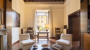 Amplia habitación con techo de madera, chimenea, dos sillones blancos, una mesa de madera con lámpara y vistas al exterior por una puerta abierta.