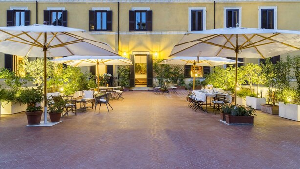 Un patio exterior con mesas y sillas, rodeado de plantas en macetas, e iluminado por luces ambientales.