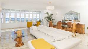 A spacious living room with a white sofa, yellow armchairs, a glass coffee table, and a wooden dining set. Large windows offer a view of the ocean. - Cubo's Apartamento Seaview Port & Free Parking / sea views / free wifi (Málaga)