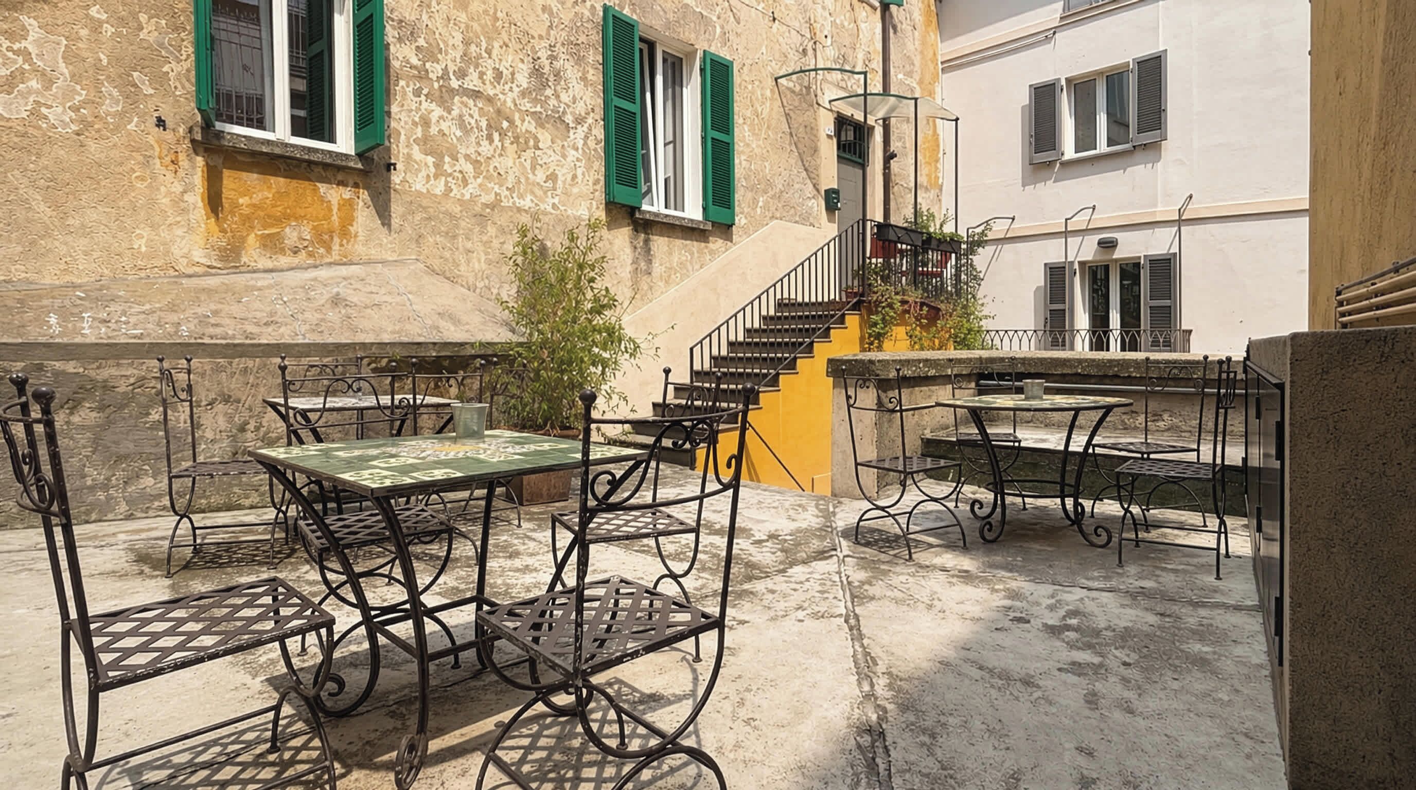 An outdoor patio area with metal tables and chairs, a staircase, and a building with stone walls and green shutters.