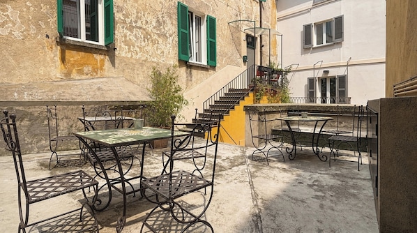 Ein Terrassenbereich im Freien mit Metalltischen und -stühlen, einer Treppe und einem Gebäude mit Steinmauern und grünen Fensterläden.