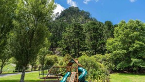 A playground with a slide and swing set, surrounded by trees and a mountain in the background. - Lakeview Oasis - Lake Whakamaru Retreat (Mangakino)