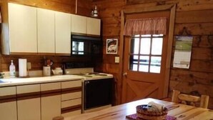 L’intérieur d’un chalet en bois comprend une cuisine avec des meubles blancs, un four à micro-ondes et une cuisinière. On y trouve également une table à manger en bois avec des bancs, ainsi qu’une fenêtre ornée d’un rideau à carreaux.